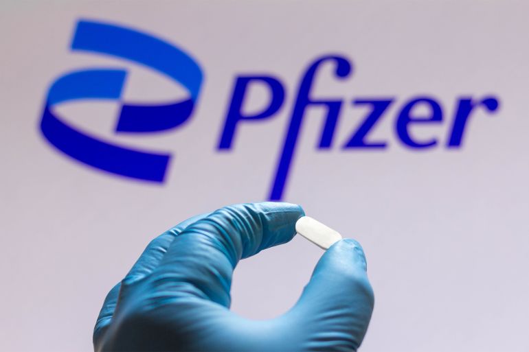 In this photo illustration Pfizer logo is seen on a screen and a hand holding a pill; Shutterstock